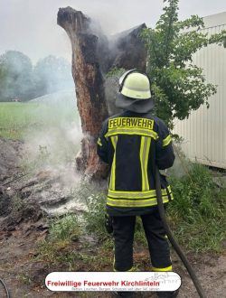 Ein Alter Baumstamm brennt und wird von einem Feuerwehrmann abgelöscht